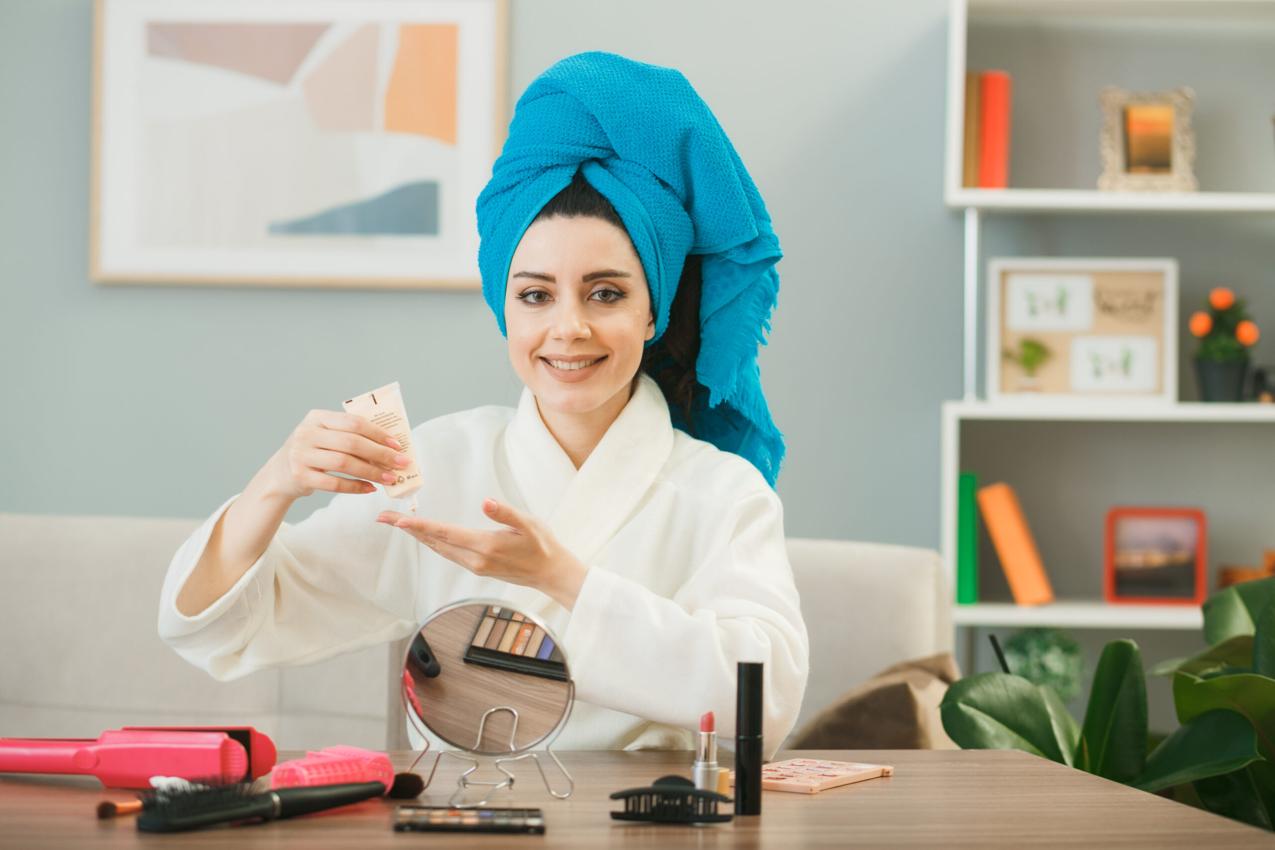 smiling looking camera young girl holding tone-up cream wrapped hair in towel sitting at table with makeup tools in living room