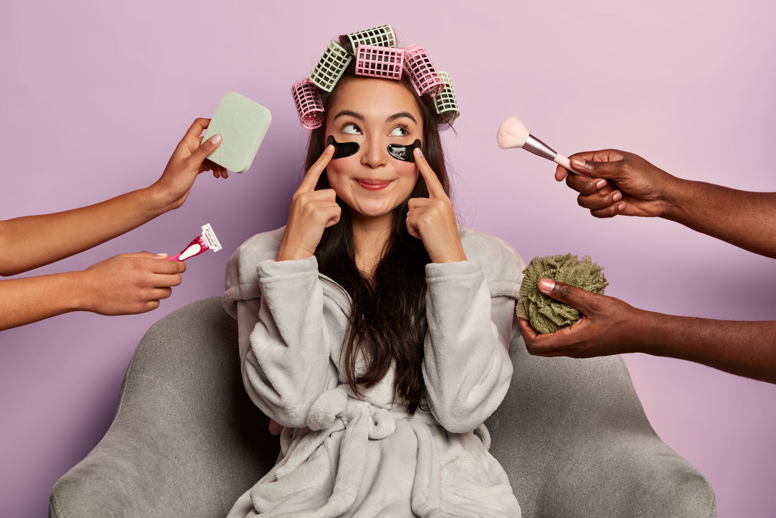 Pretty Korean woman points at eye patches, wears bathrobe, sits at home after shower, has healthy skin and complexion, isolated over purple wall. Beauty industry, cosmetology, skin care concept